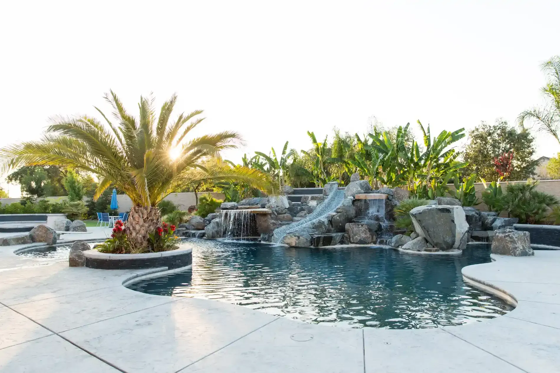 Resort-style pool with rock waterfall, palm trees, and clear water at sunset