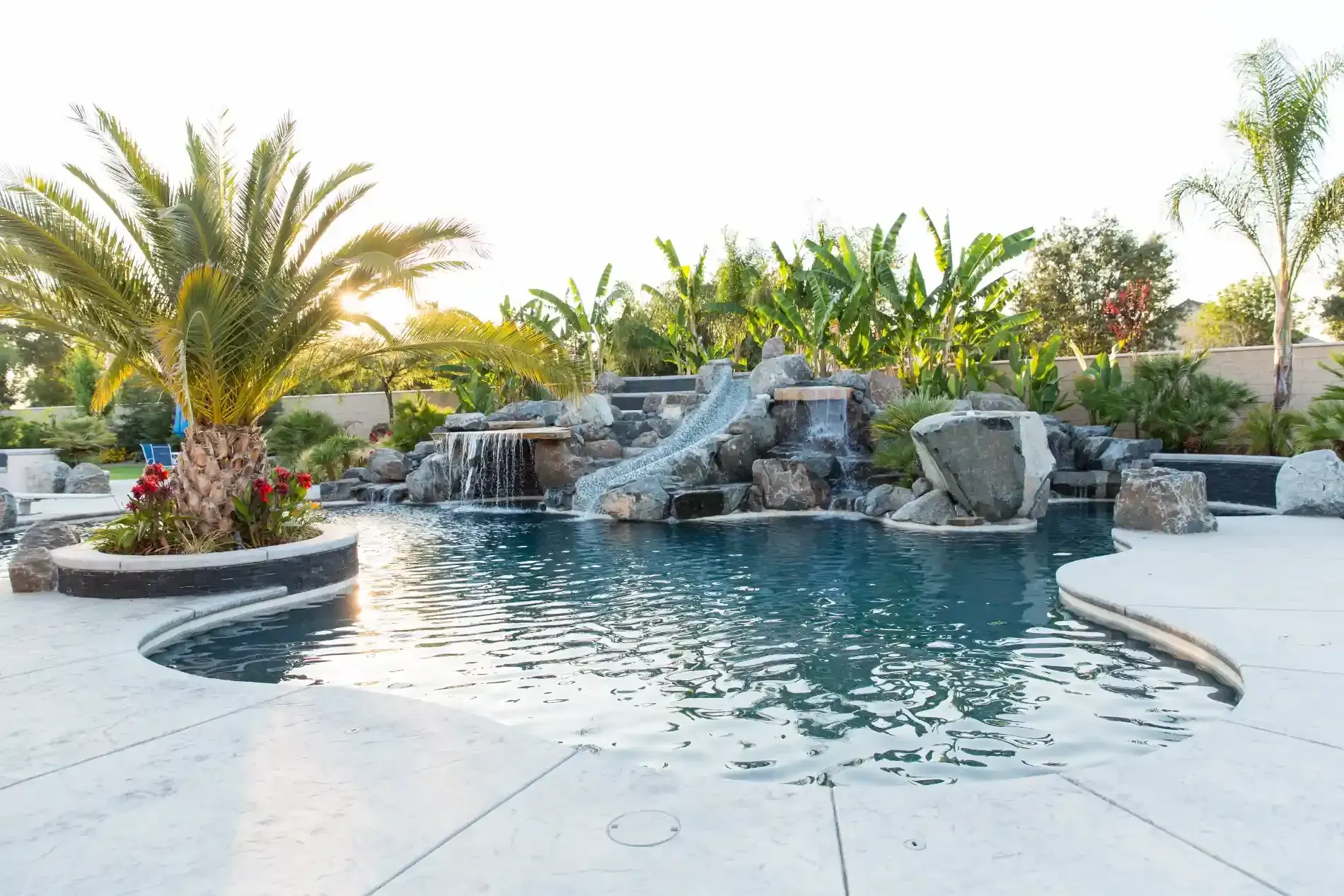 Backyard pool with rock waterfall, palm trees, and white stone deck in bright daylight