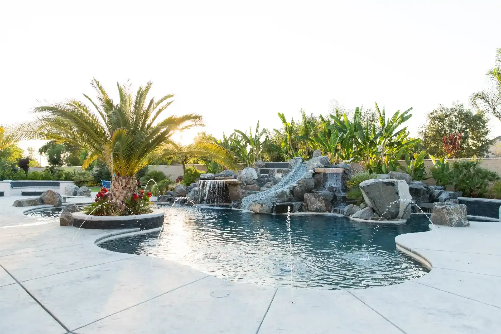 Tropical resort pool with rock waterfall, palm trees, and lounge chairs at sunset