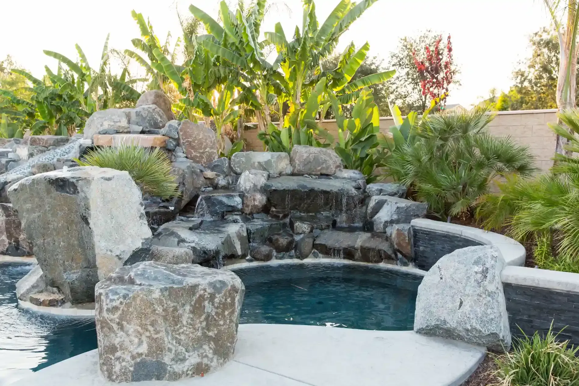 Backyard rock waterfall cascading into a small pool with tropical plants and stone seating area
