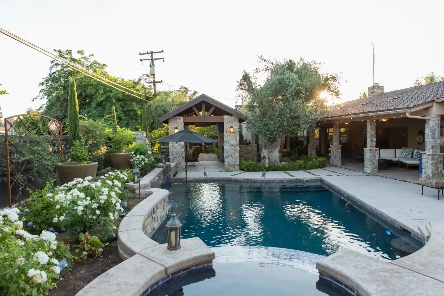 Backyard pool with stone patio, flowers, and a shaded pavilion at sunset