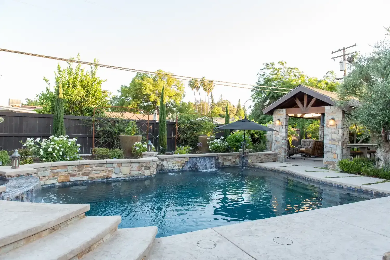Stone backyard pool with blue water, steps, and a shaded gazebo beside a stone wall.