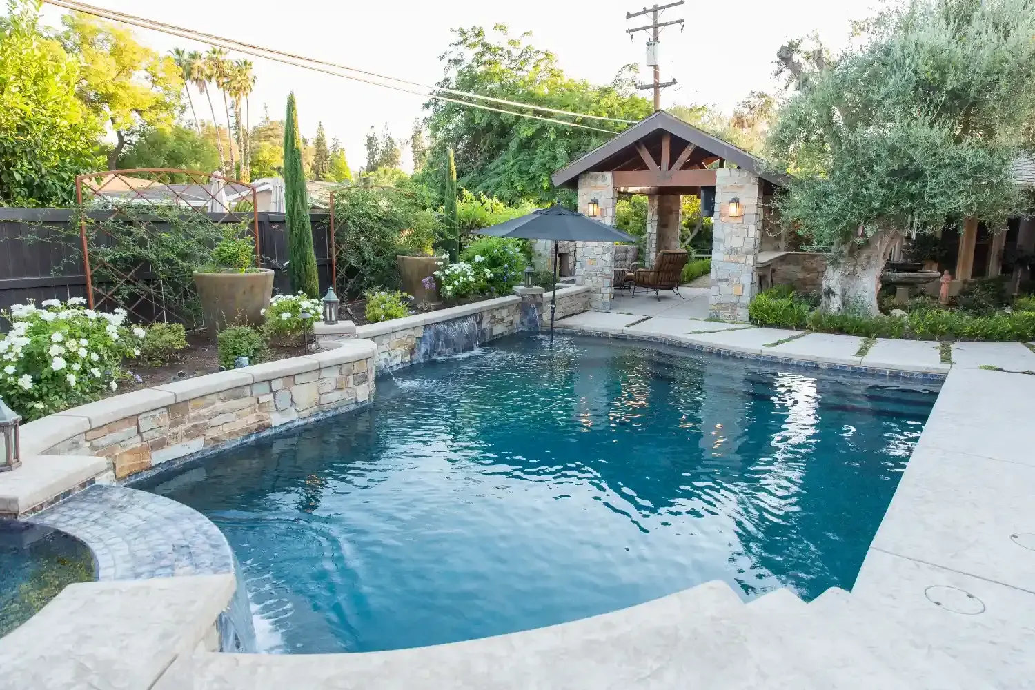 Backyard swimming pool with stone surround, garden landscaping, and a covered pavilion at the far end