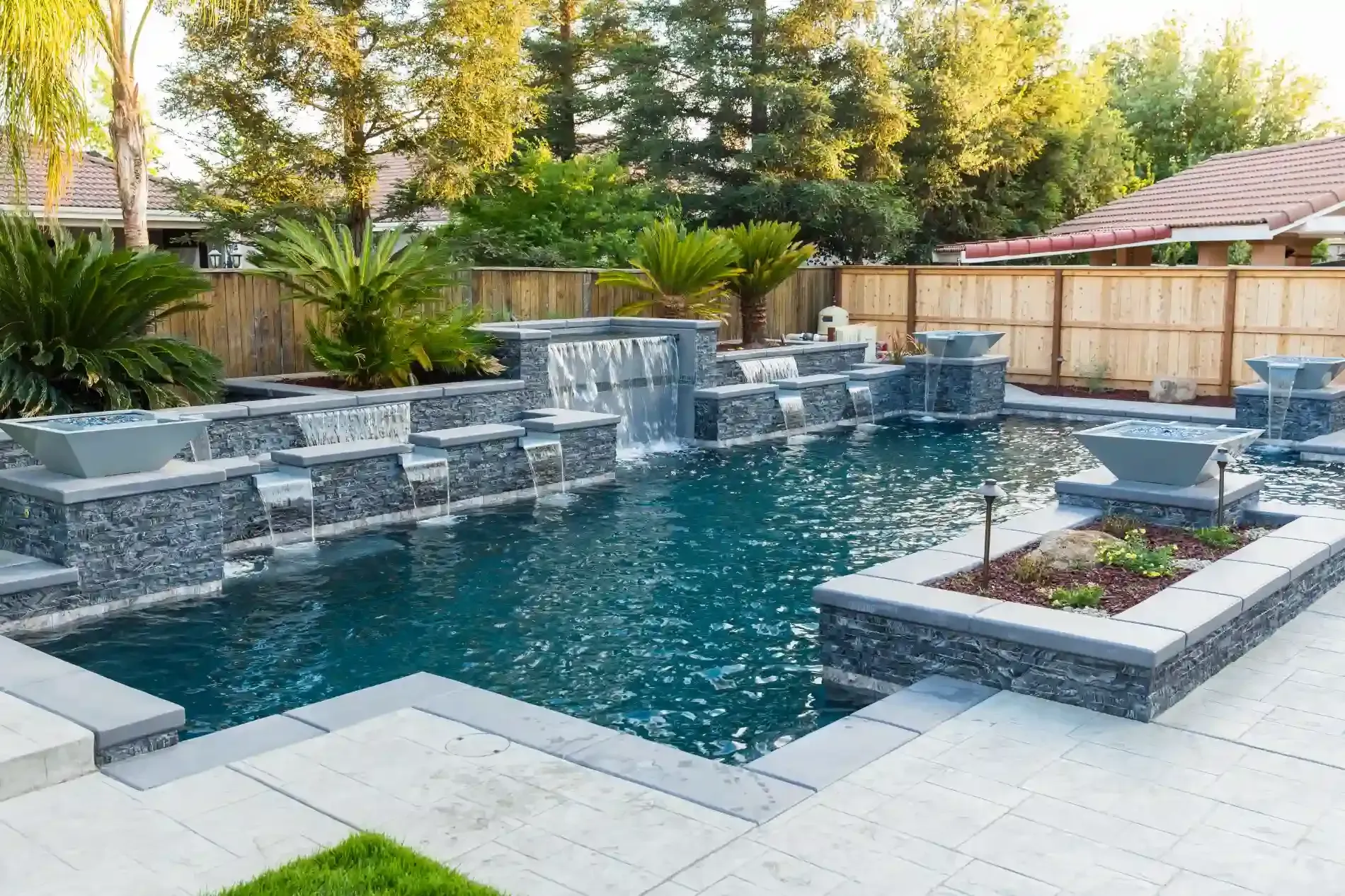 Modern backyard pool with stone waterfall, raised spa, and lounge chairs beside a wooden fence