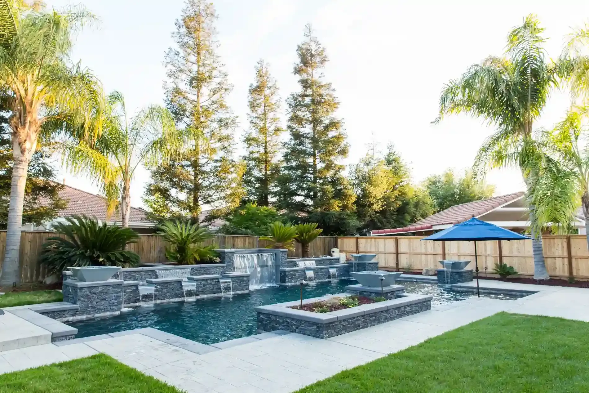 Backyard patio with a modern stone fire pit, pool, palm trees, and lounge chairs
