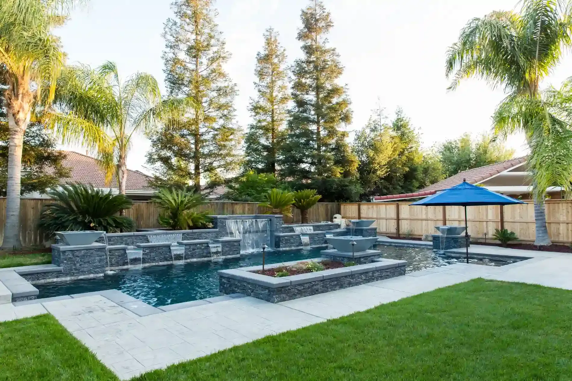 Modern backyard with a stone pool, fire feature, green lawn, and tall trees at sunset