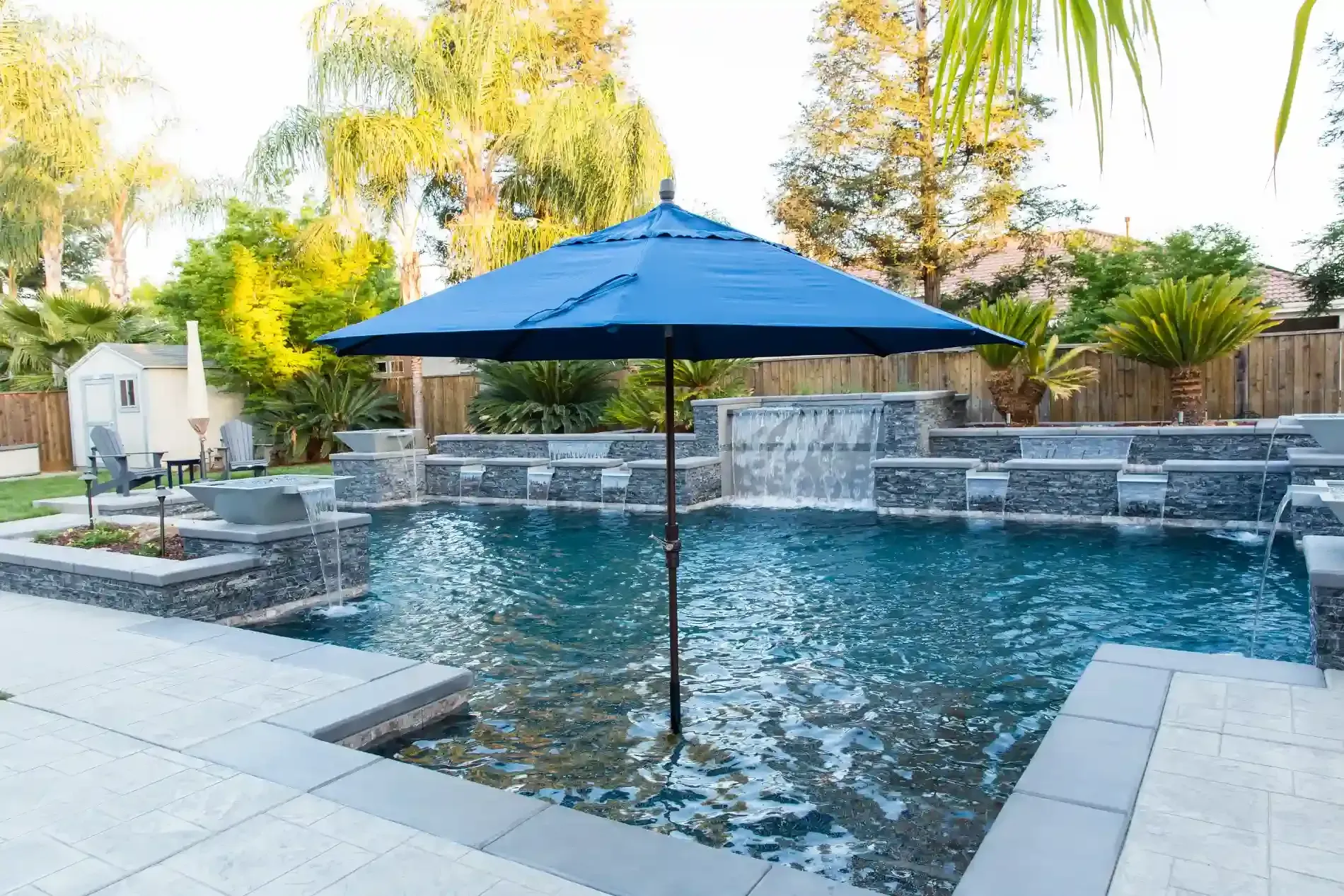 Backyard pool with a blue umbrella and stone waterfall under palm trees