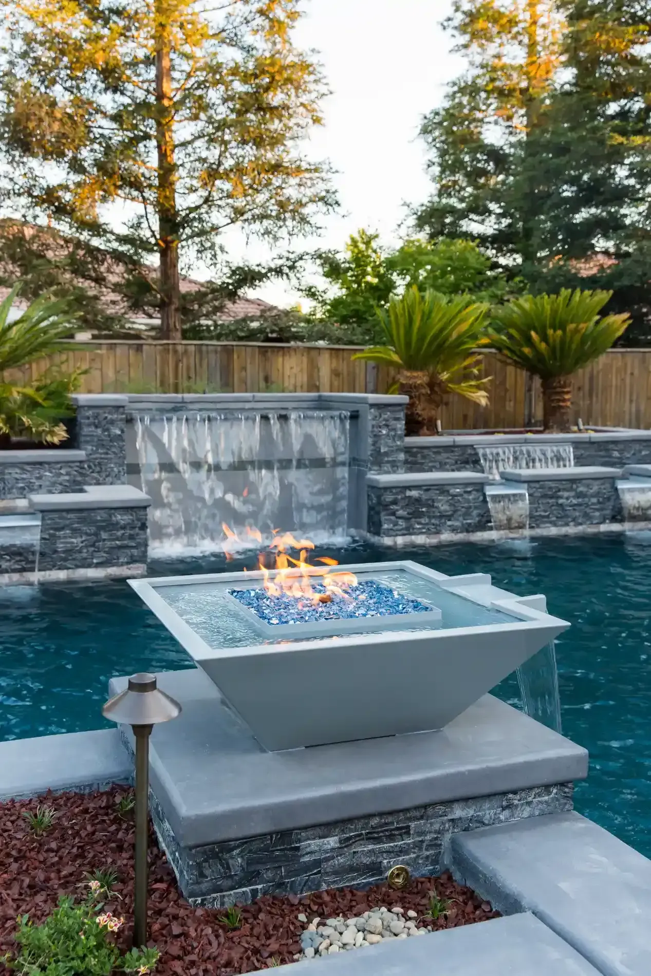 Backyard pool with a modern fire bowl fountain and stone waterfall, framed by autumn trees