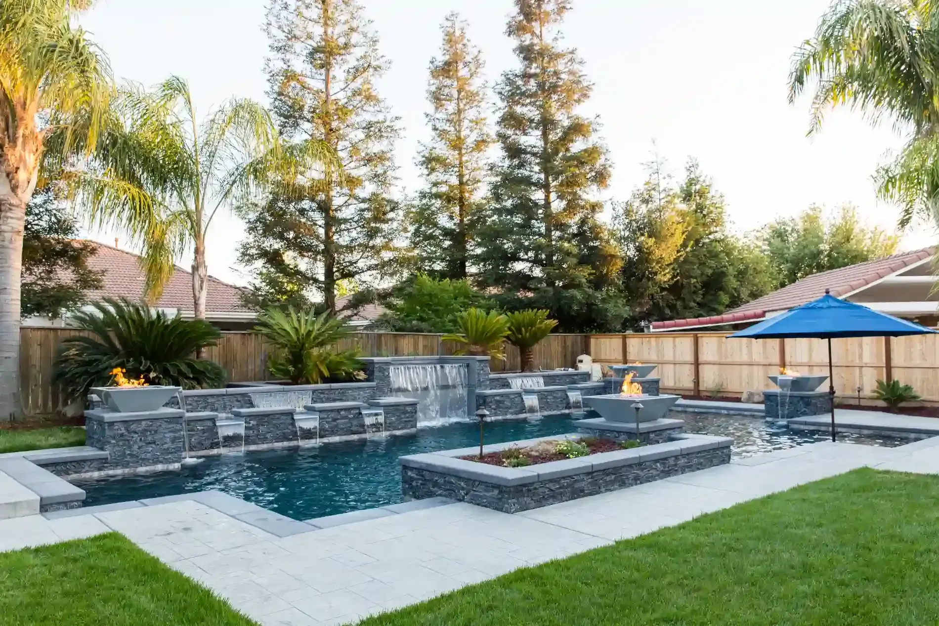 Backyard pool with gray stone ledges, blue water, and a lawn bordered by tall trees and umbrellas