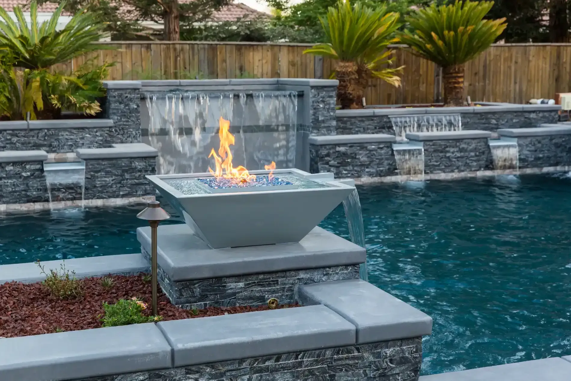 Backyard pool with a modern fire bowl on a stone platform and waterfall features, lit with a small flame.
