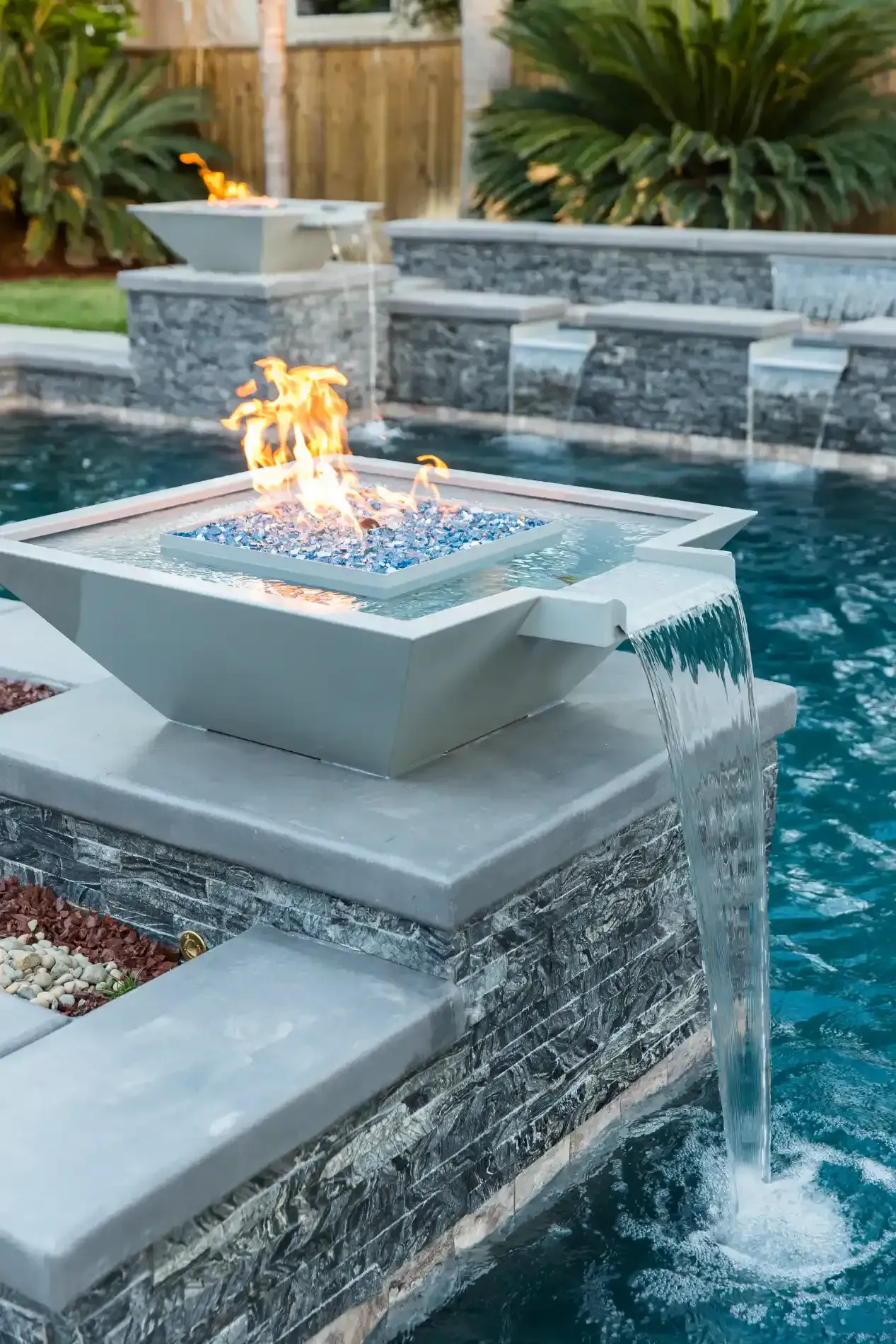 Modern backyard fire feature beside a pool, with water cascading over stone ledges
