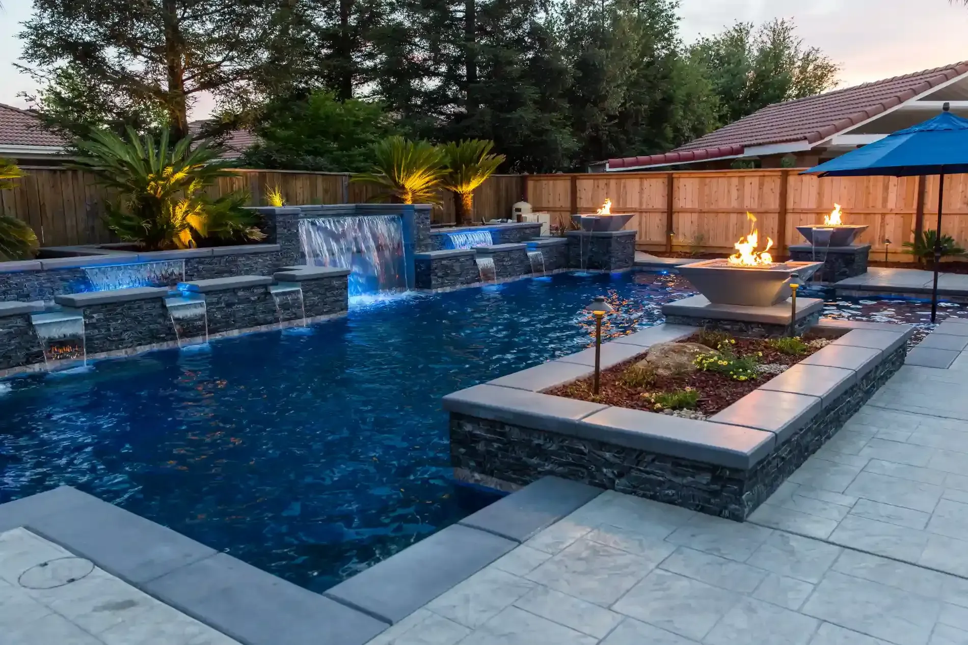 Modern backyard pool with waterfall, fire feature, and stone patio at dusk