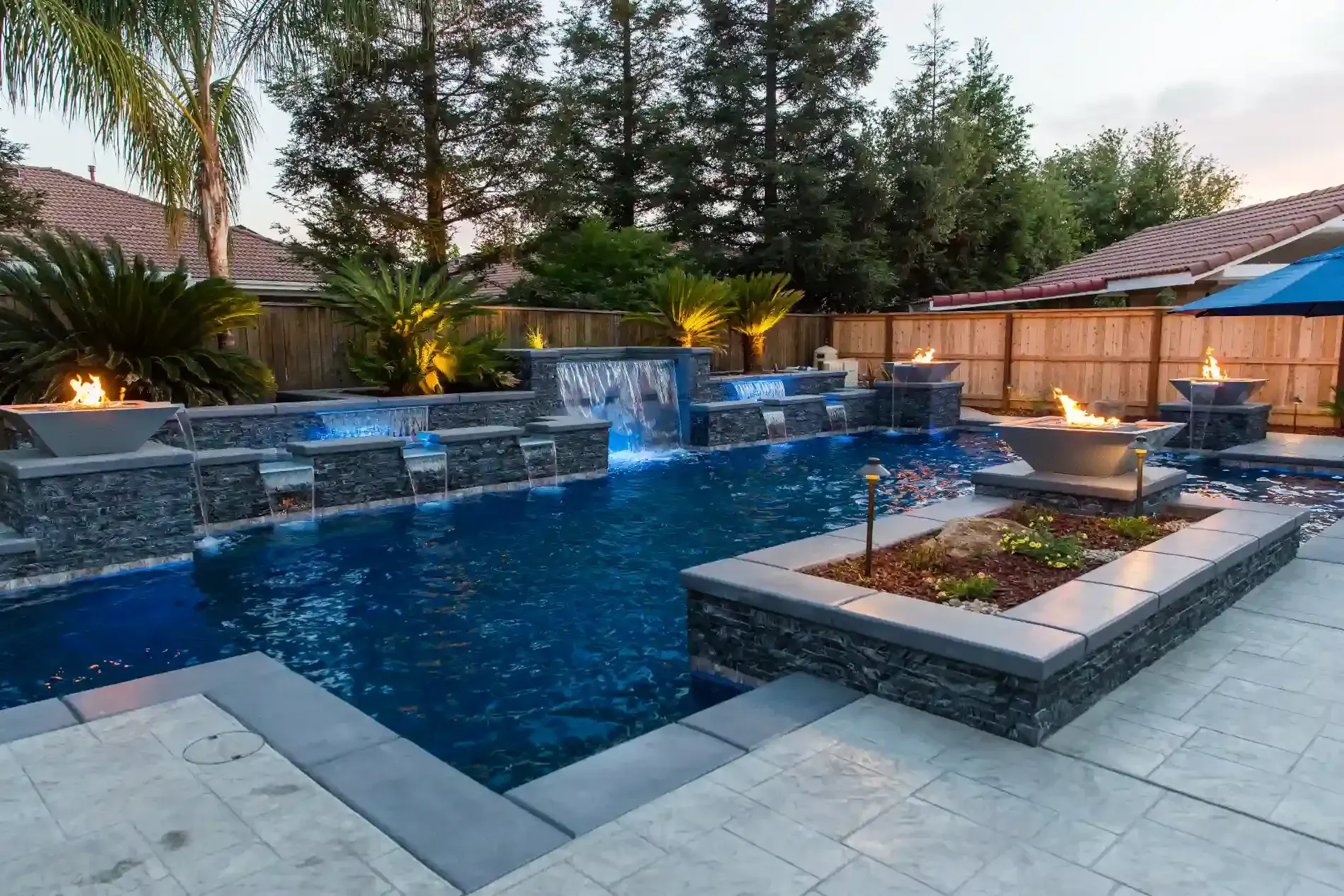 Modern backyard pool with stone water features, glowing blue lights, and a raised planter at dusk
