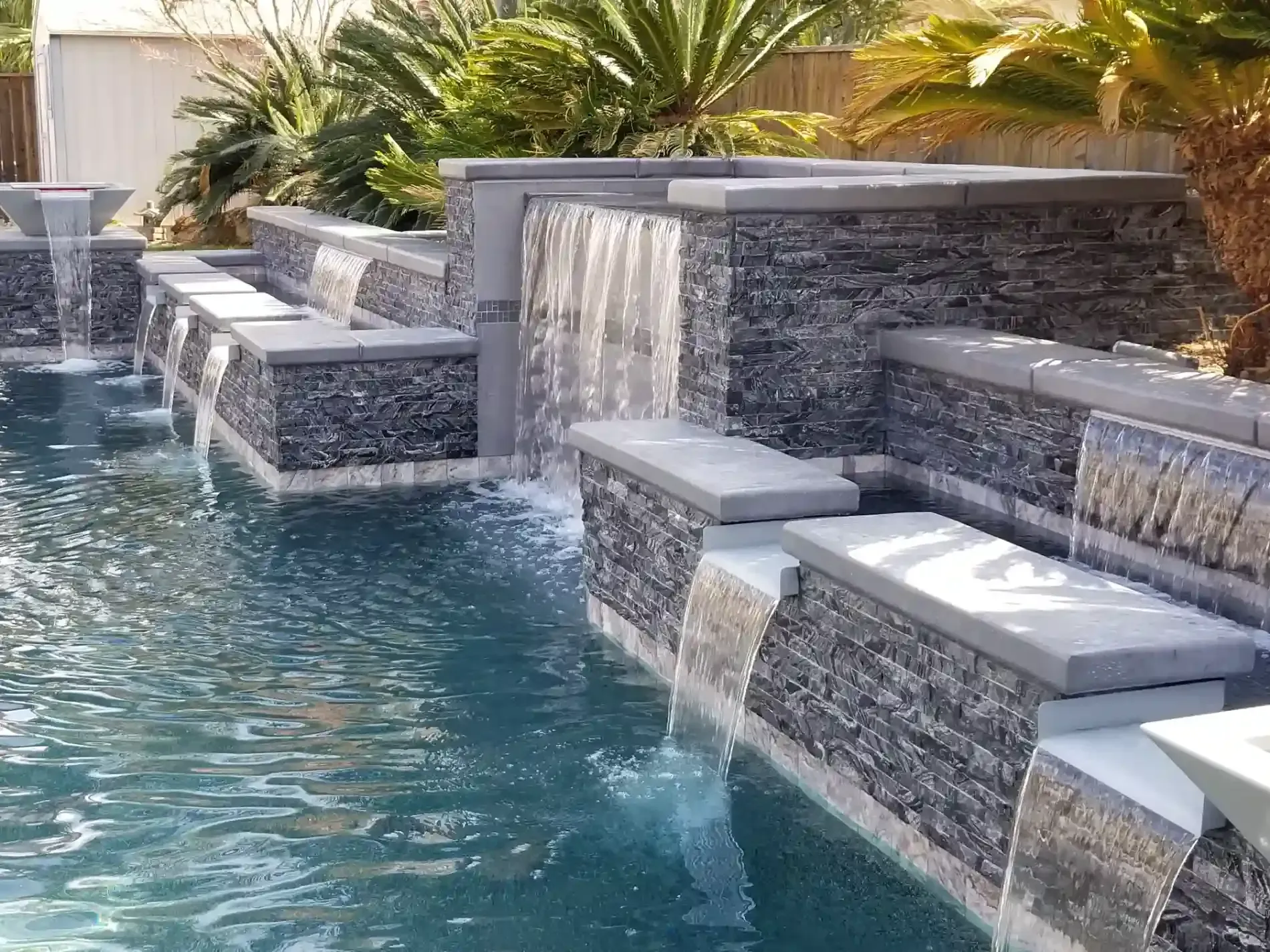 Luxury backyard pool with tiered waterfalls, stone seating, and palm plants