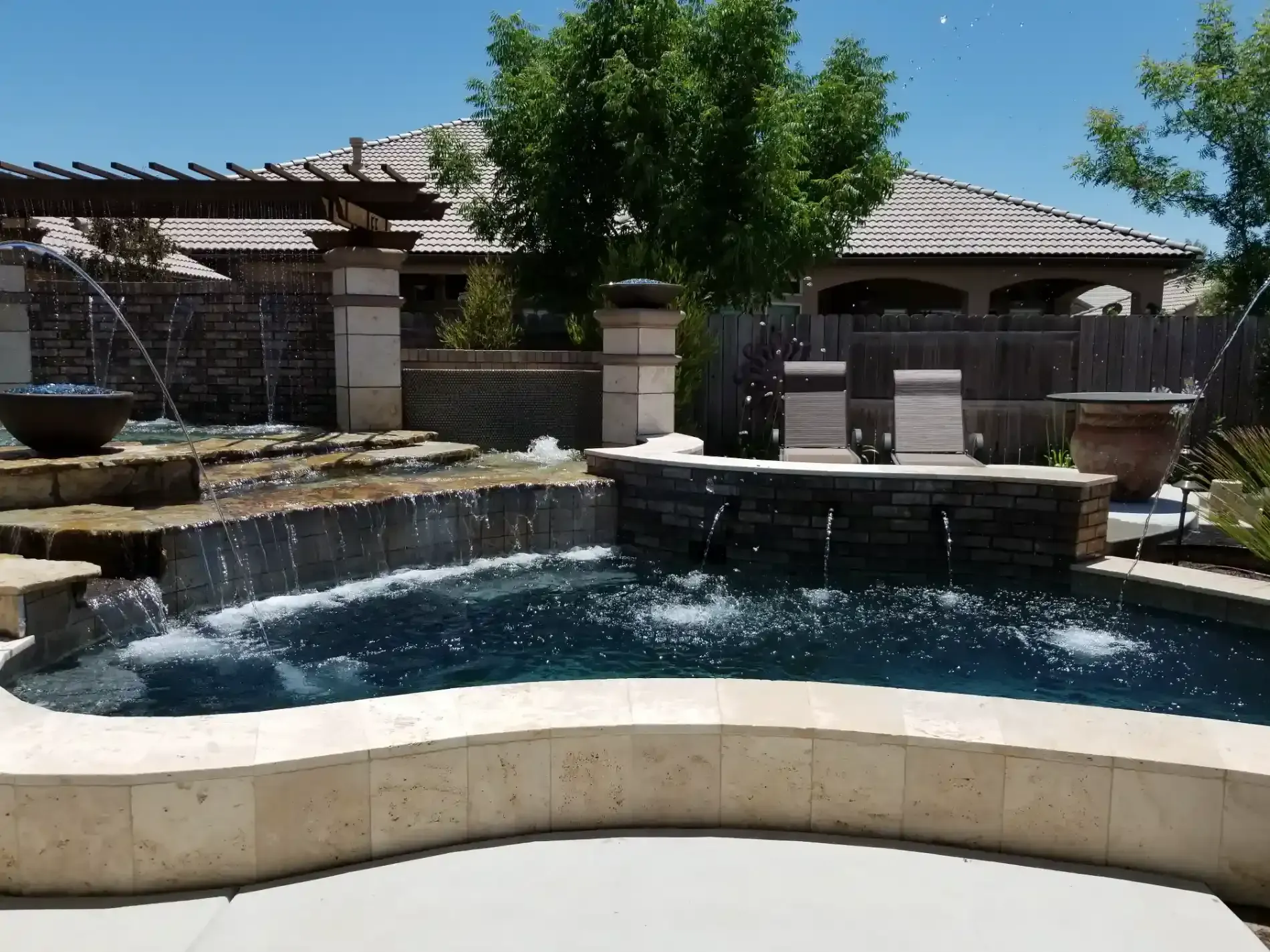 Backyard pool with stone waterfall, patio seating, and a large tree under a clear blue sky