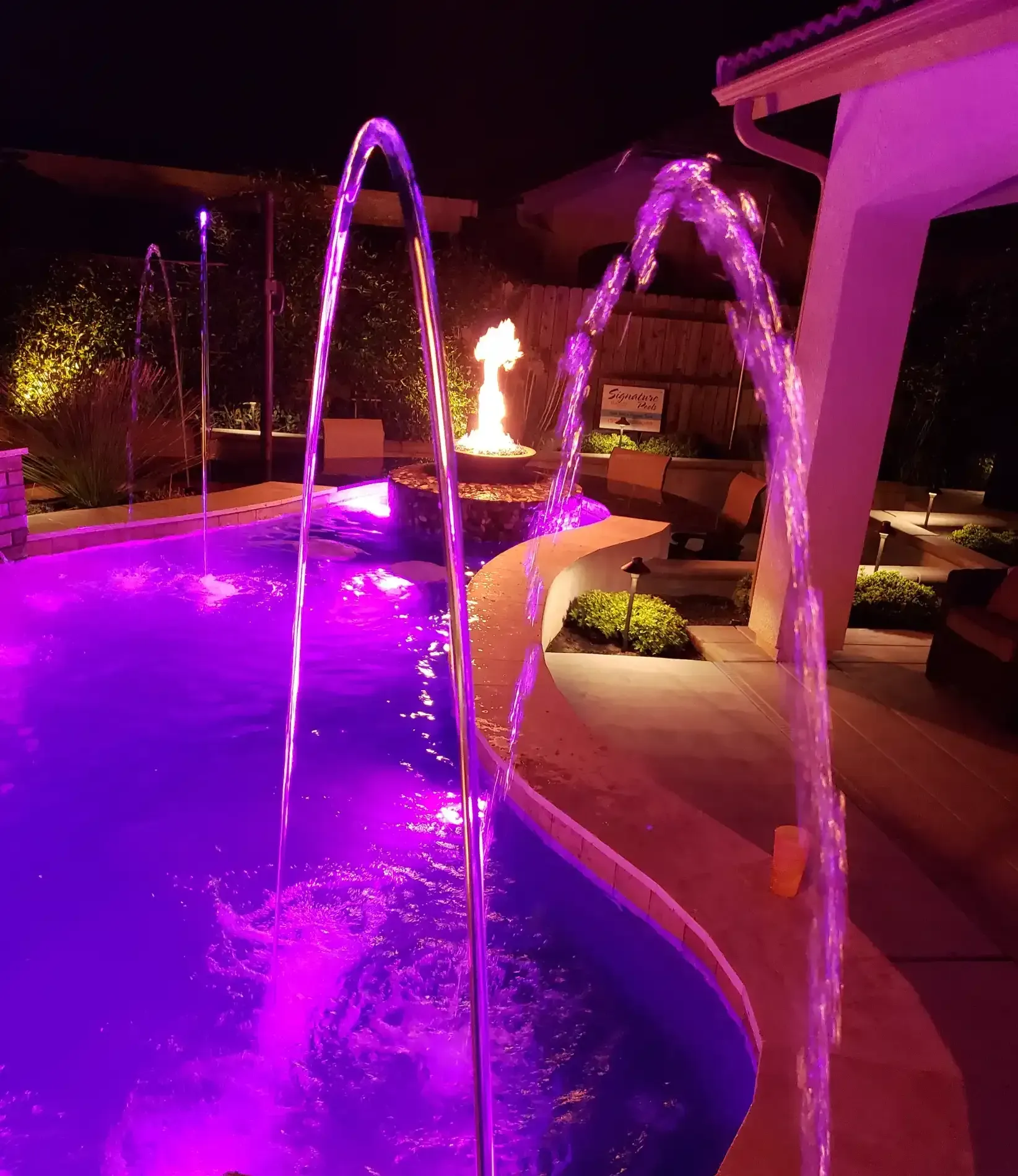 Purple-lit backyard pool with arcing water jets and a fire feature at night