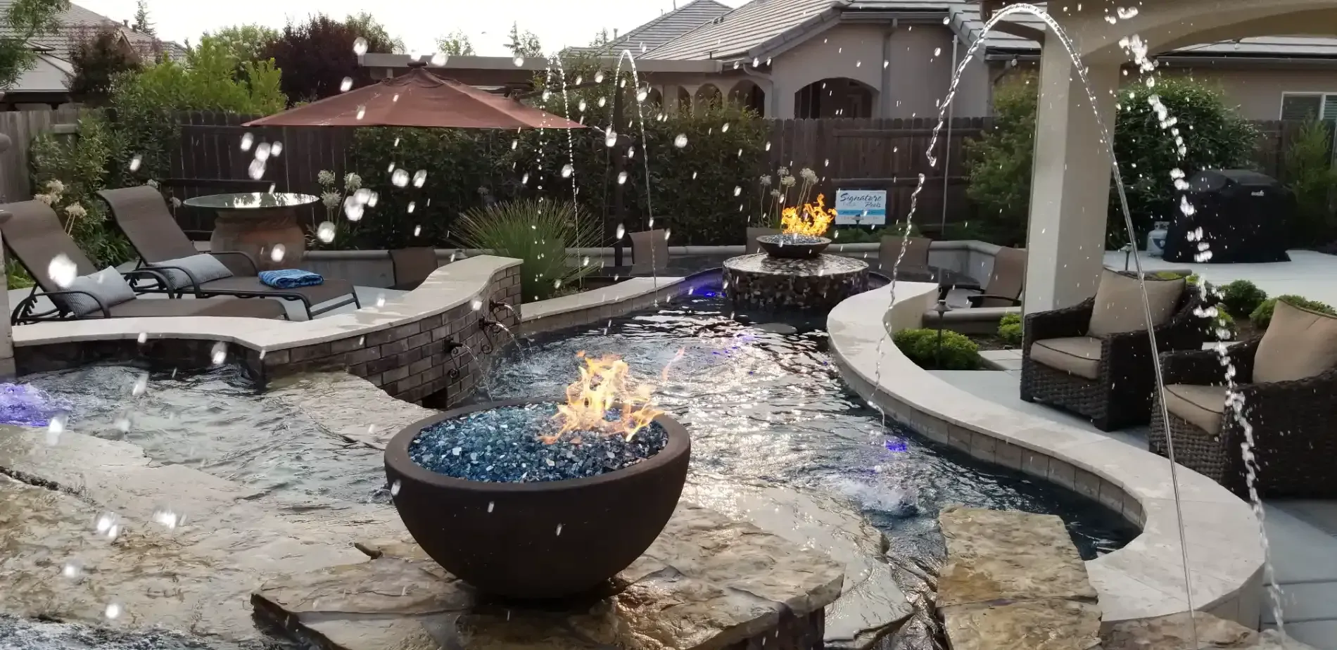 Backyard patio with fire bowls, stone pathways, lounge chairs, and a lit fire pit at dusk.