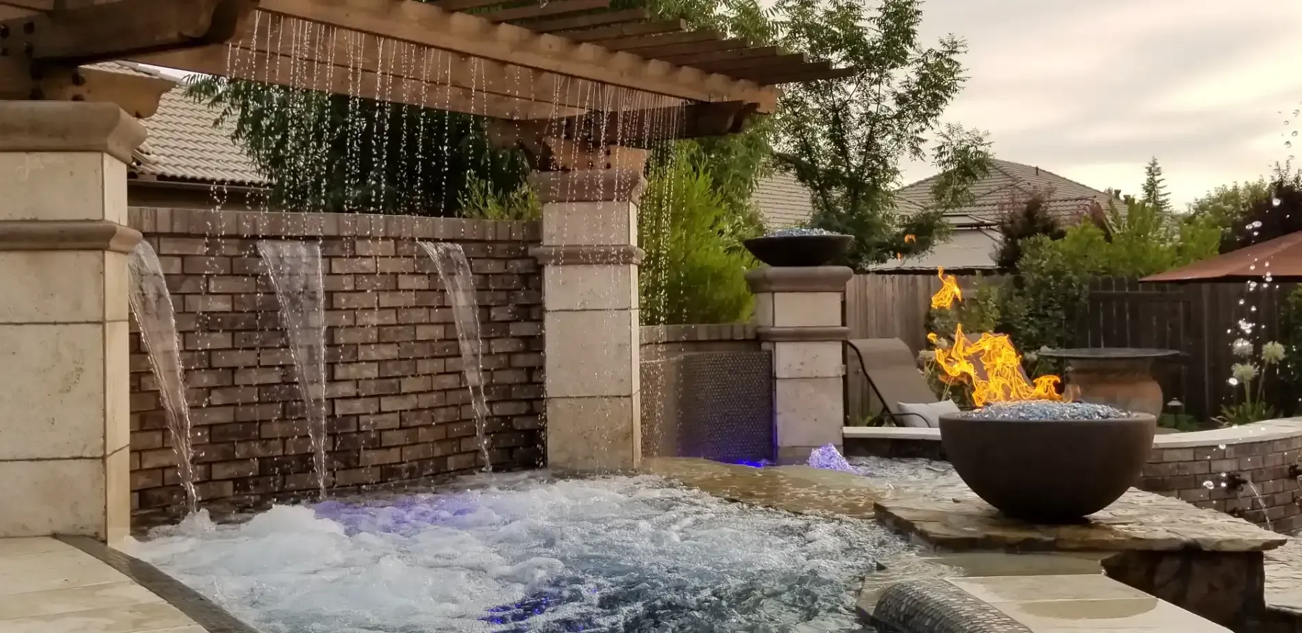 Backyard waterfall with stone wall, bubbling water, and a fire bowl beside a patio pool