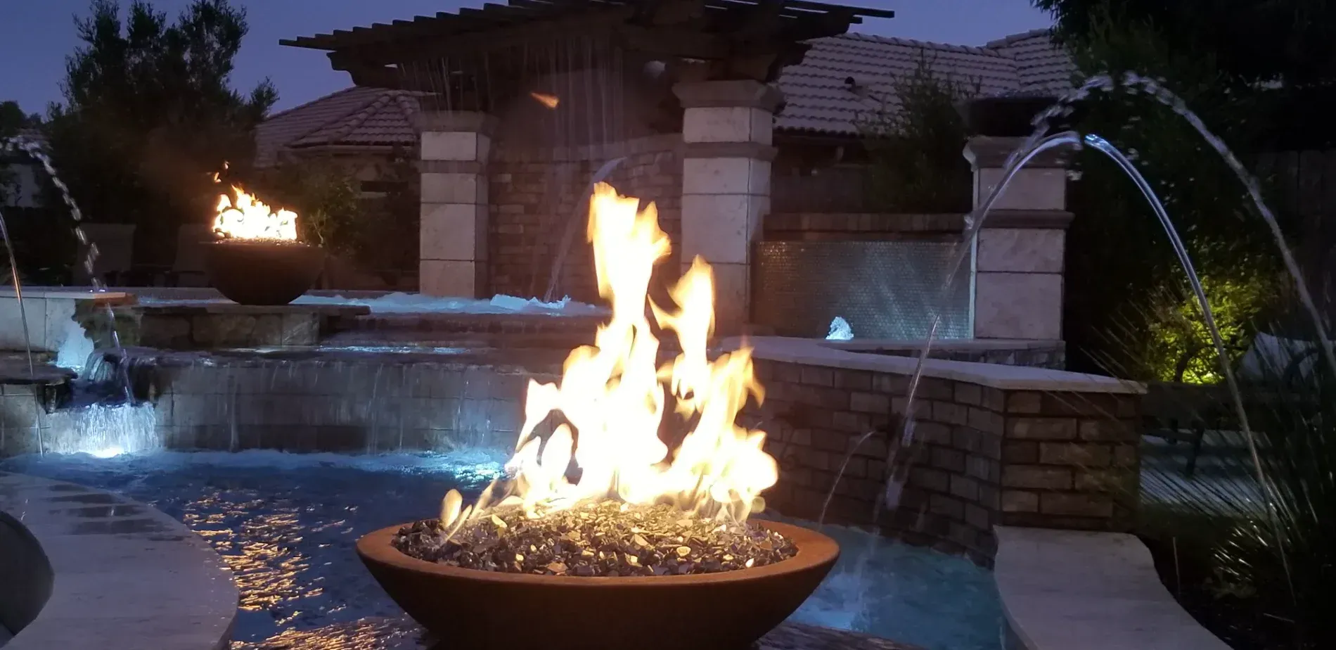 Backyard pool at dusk with a fire bowl in the foreground and lit water jets in the background