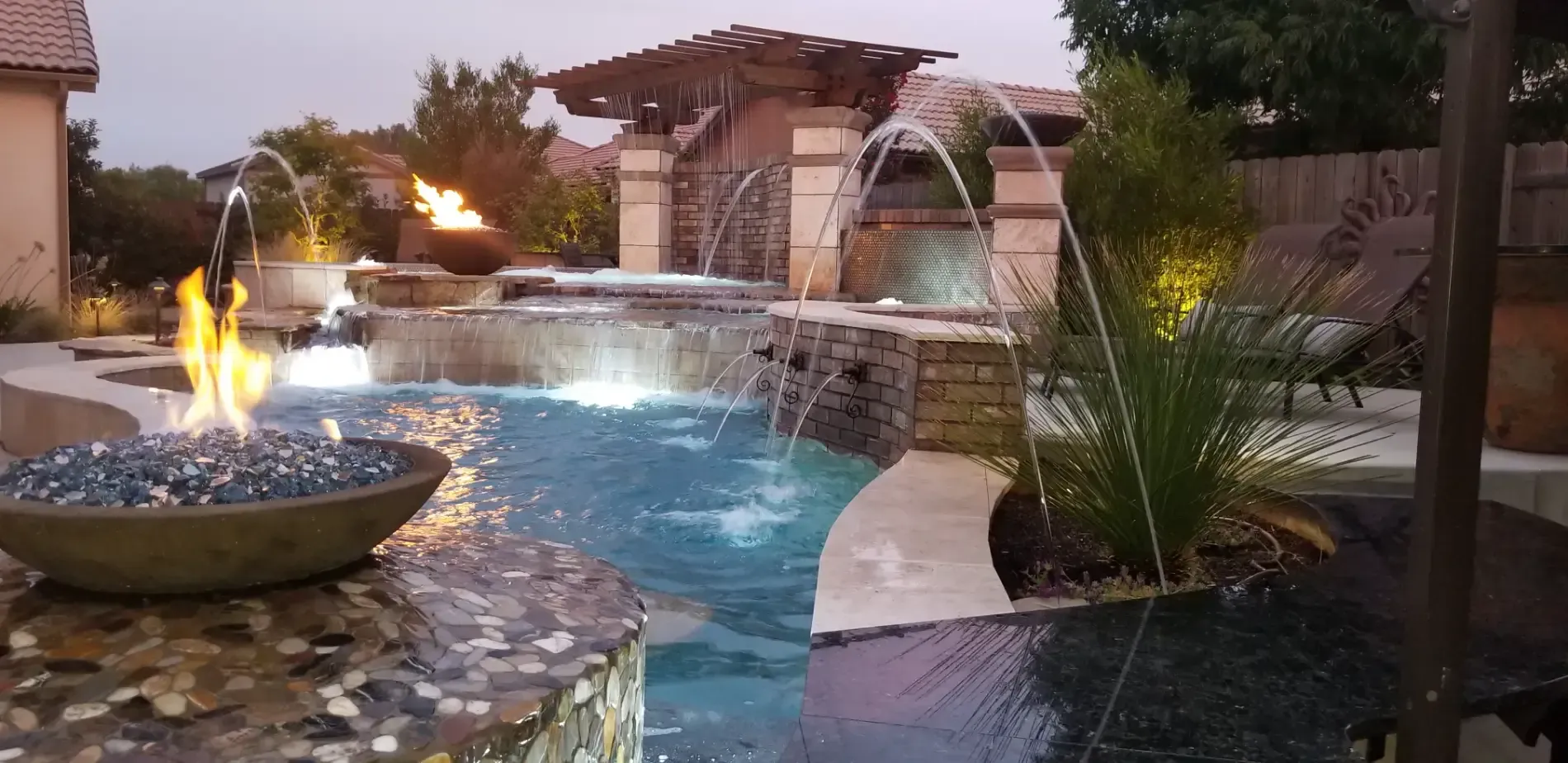 Backyard pool with stone waterfall, fire feature, and tropical landscaping at dusk