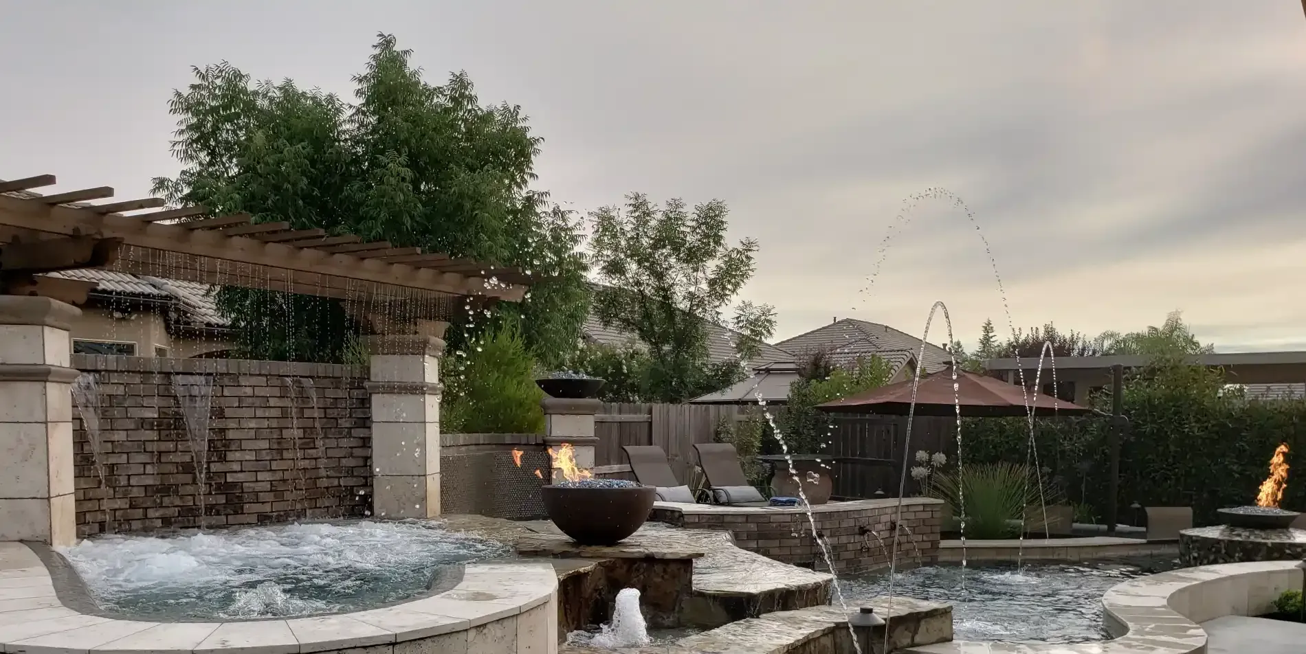 Backyard pool with stone waterfall and covered pergola beside a landscaped patio at dusk