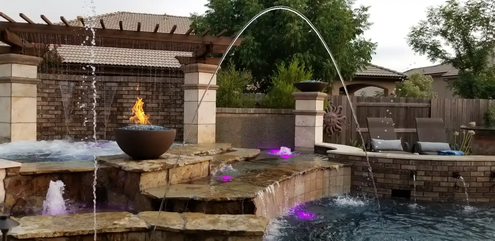 Stone backyard fountain with arched water jets and a lit fire bowl on a patio