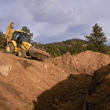 Yellow backhoe digging in a brown dirt hillside, with green trees and a cloudy sky in the background.
