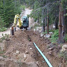 An excavator digging a trench in a forest, laying a blue pipe.