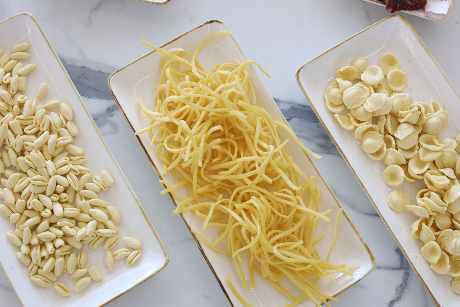 Tre piatti rettangolari con diversi formati di pasta: orzo, tagliolini e orecchiette, disposti su una superficie bianca.