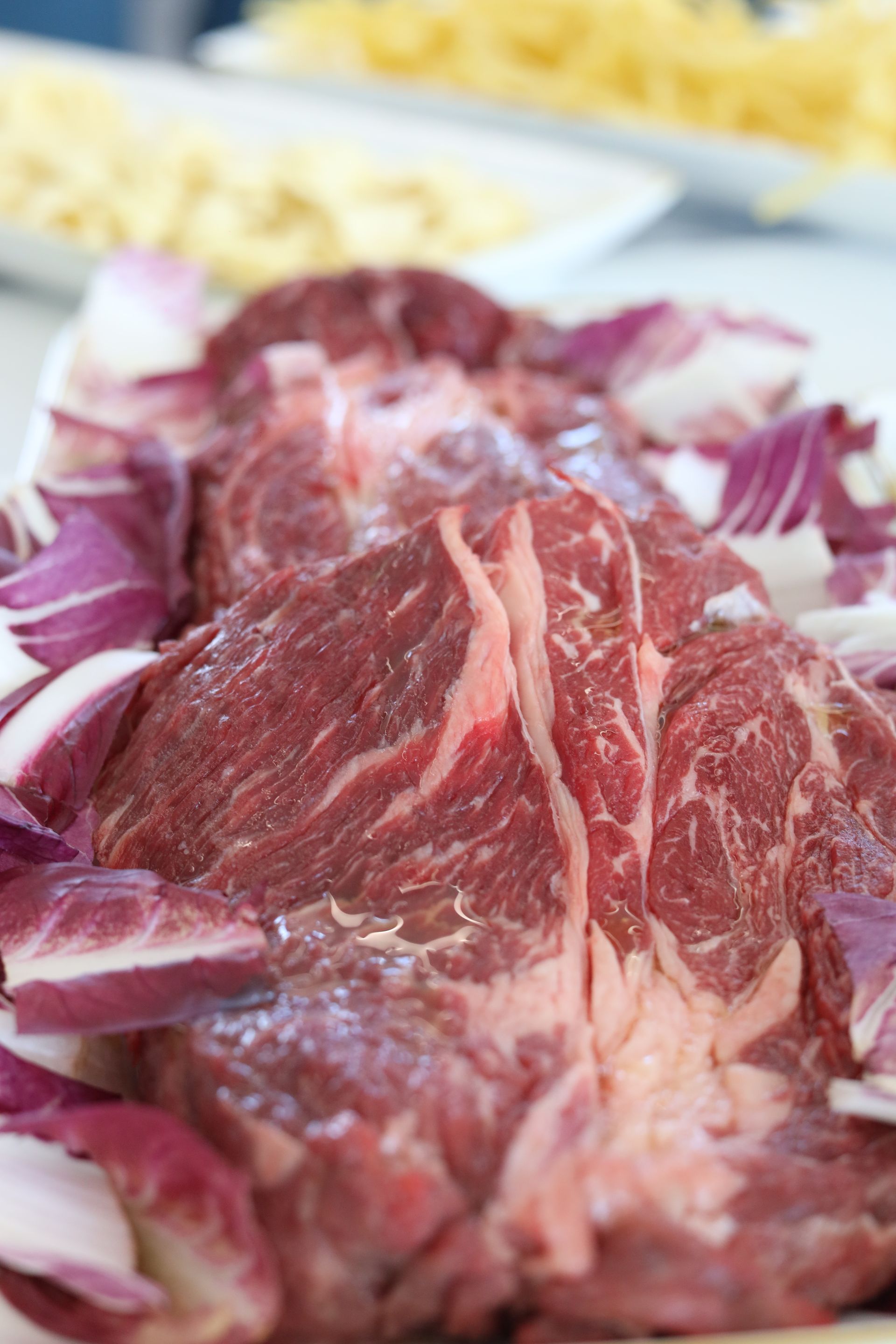 Primo piano di bistecche di manzo crude su un piatto, circondate da foglie di radicchio rosso, con piatti bianchi sfocati sullo sfondo.