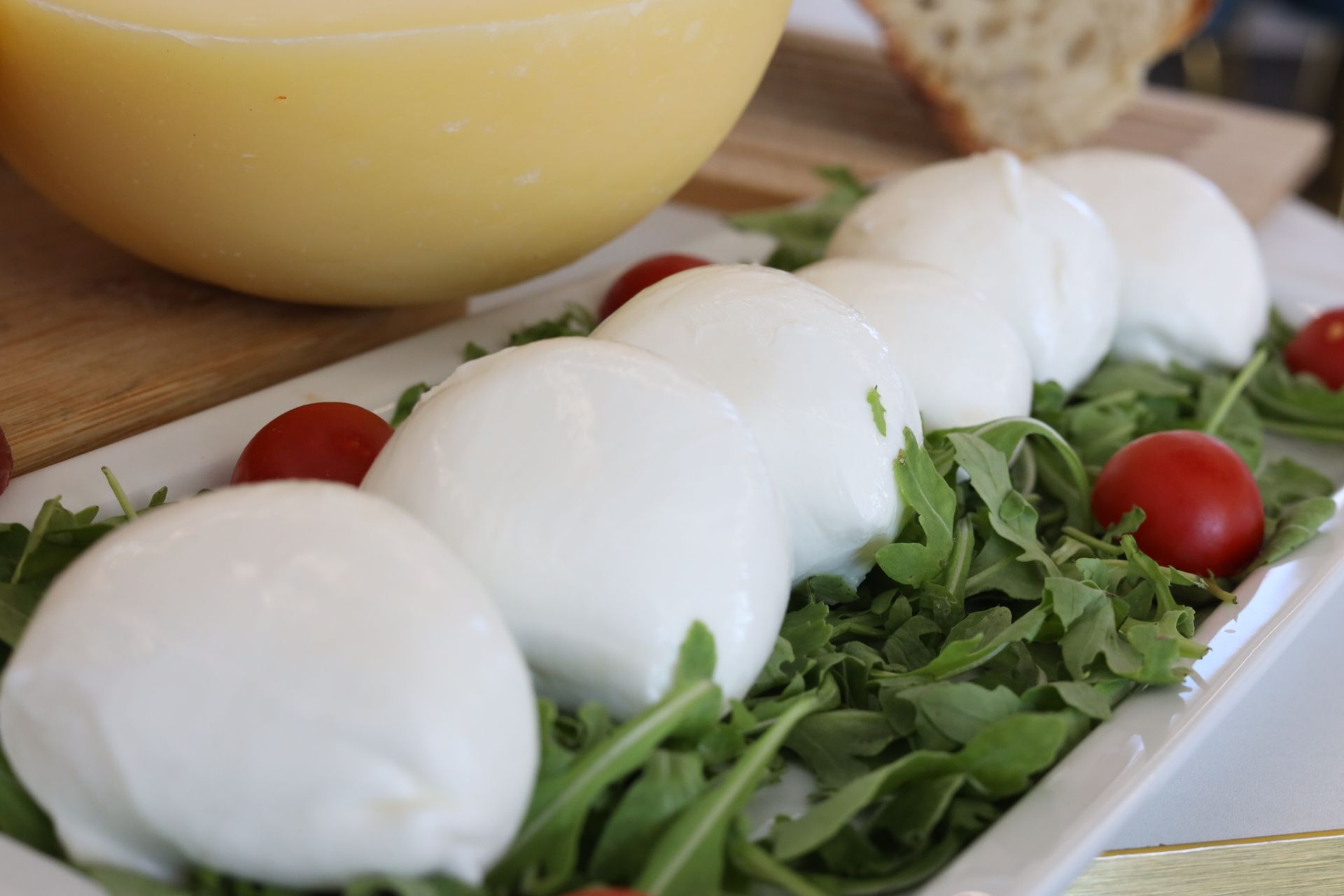Mozzarella fresca su un letto di rucola, guarnita con pomodorini. Sullo sfondo, una grande forma di formaggio e del pane.