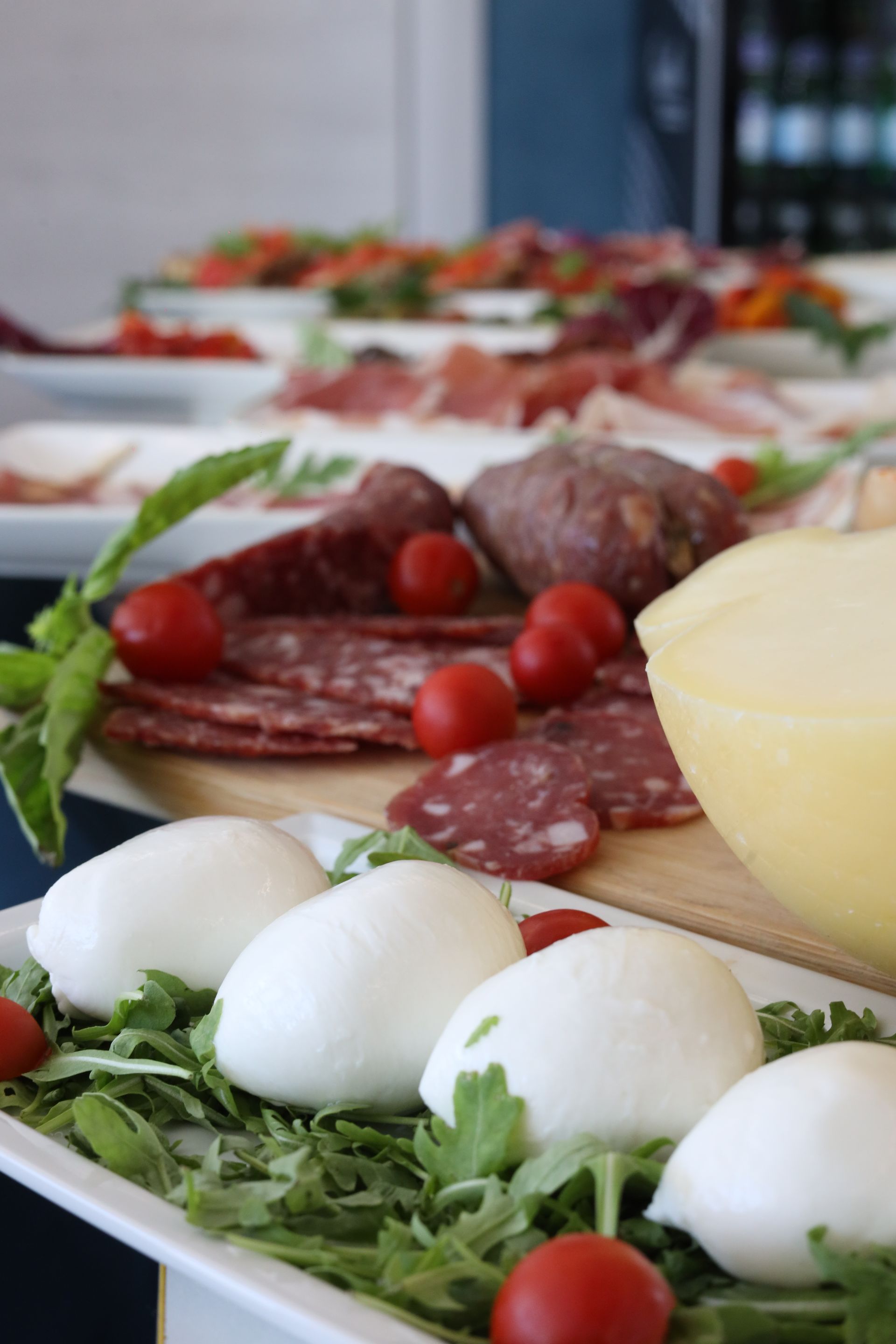 Un piatto di salumi, formaggi e pomodori italiani è disposto su un tavolo bianco. Sullo sfondo, altri piatti con pietanze simili.