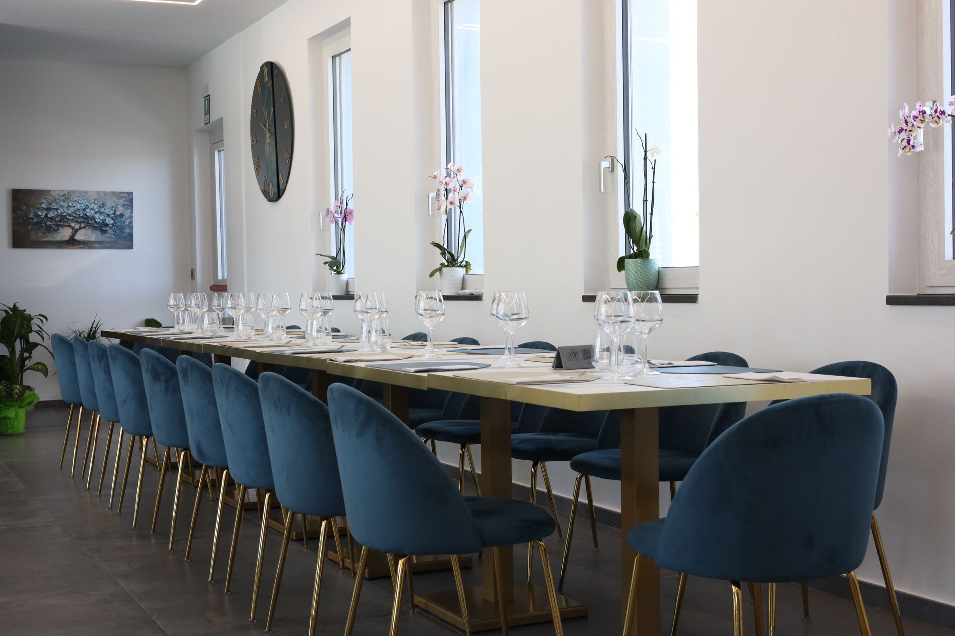 Un ristorante moderno con sedie blu, tavoli bianchi e una parete decorativa color verde acqua. La luce del sole filtra attraverso una finestra sui tavoli apparecchiati per i commensali.