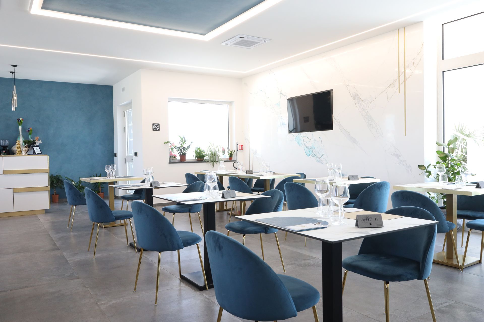 Interno di un ristorante con la tavola apparecchiata in primo piano; una parete con texture color verde acqua e un mobile contenitore bianco con piante e un sistema di cassa sullo sfondo.