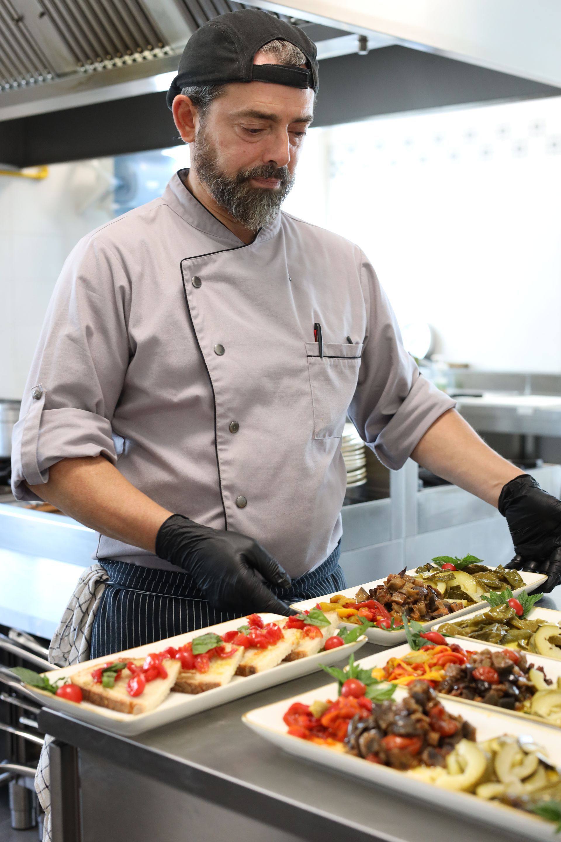 Uno chef con guanti neri mescola la pasta in padella, sullo sfondo un altro cuoco con una retina per capelli. Ambiente cucina con pentole piene di ingredienti.