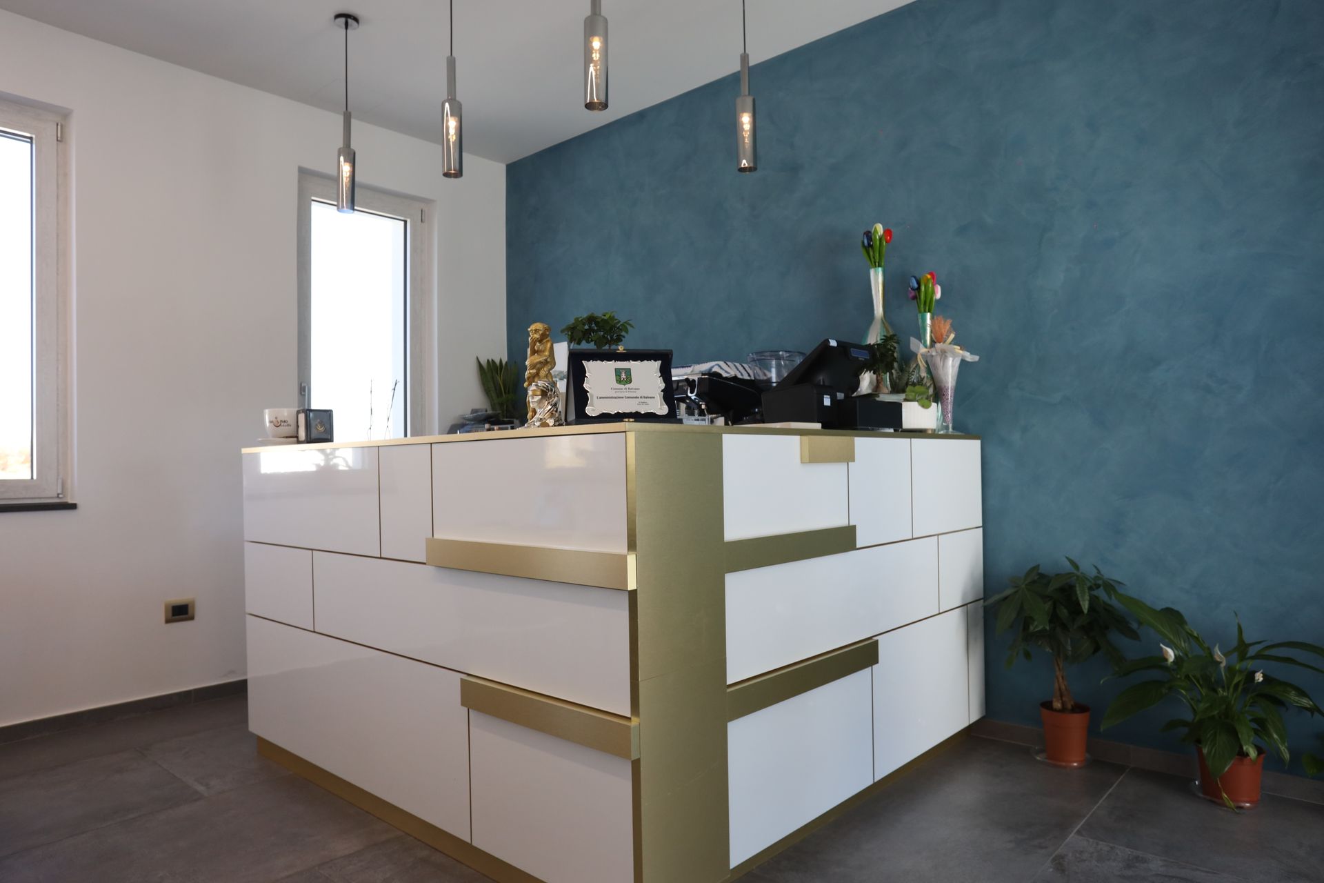 Bancone della reception bianco e oro contro una parete blu testurizzata, con luci sospese e piante in vaso.