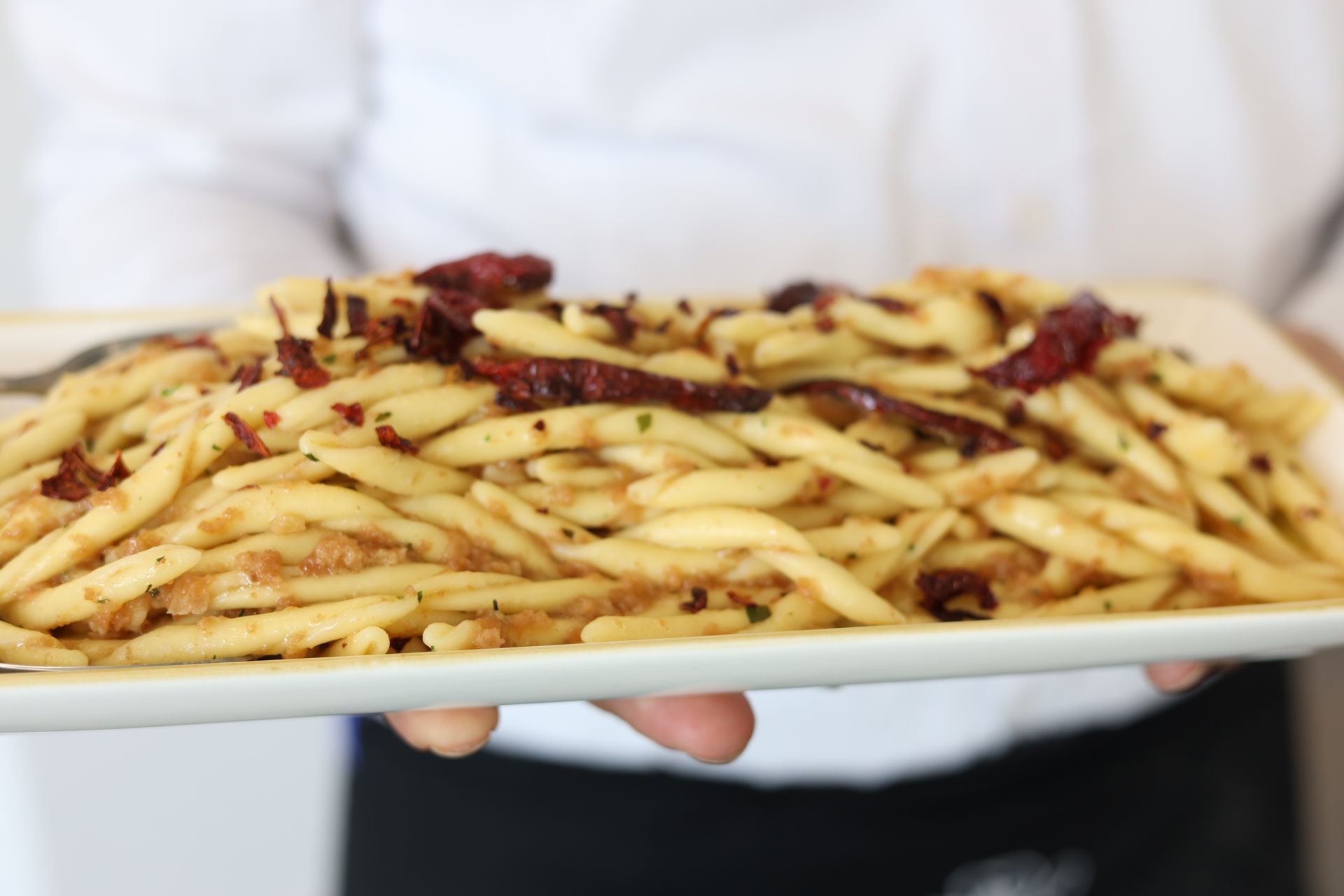 Persona che tiene in mano un piatto di pasta con peperoncino ed erbe aromatiche. La pasta è dorata e il piatto è bianco.