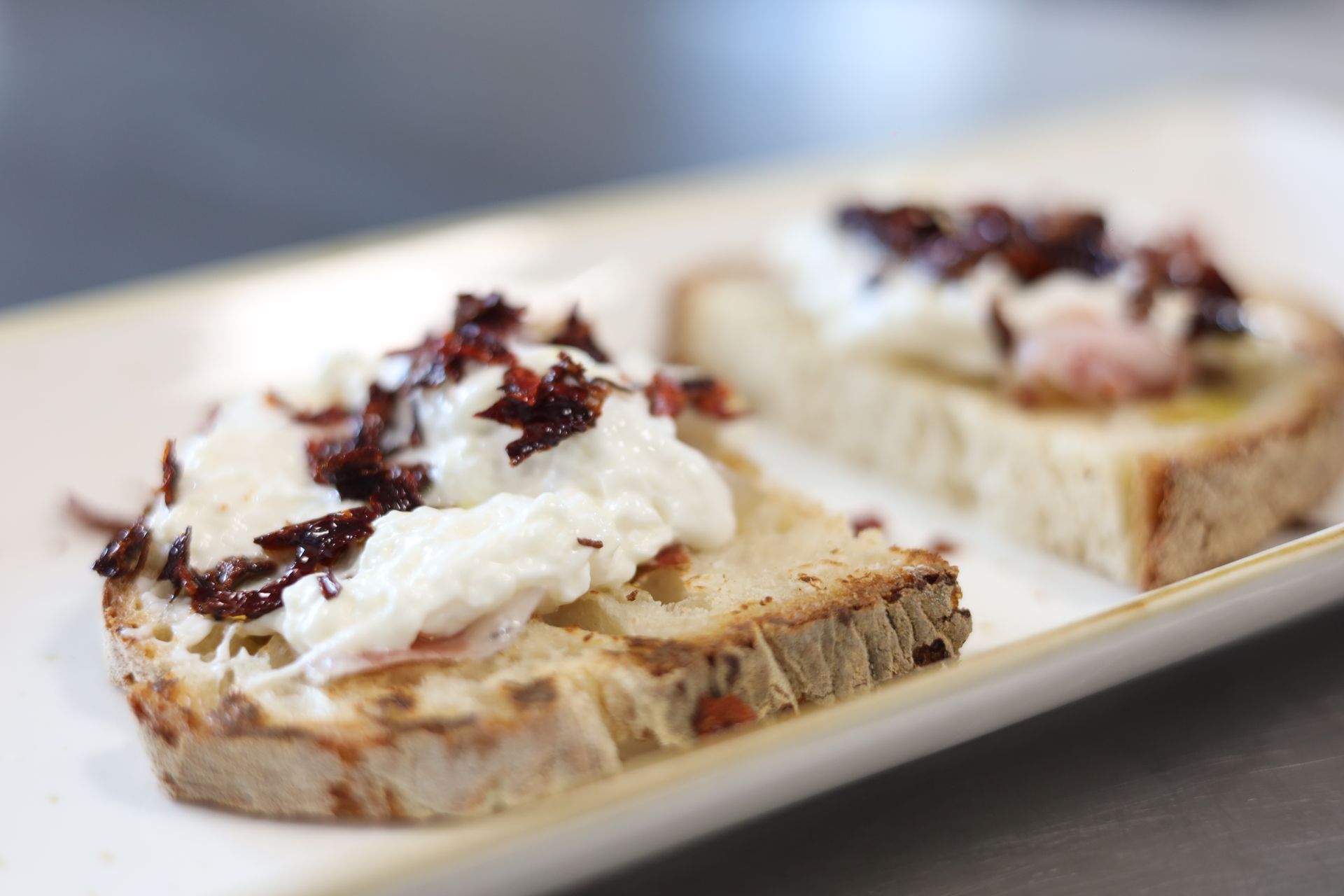 Due fette di pane tostato con formaggio bianco e ingredienti scuri tritati su un piatto rettangolare bianco.