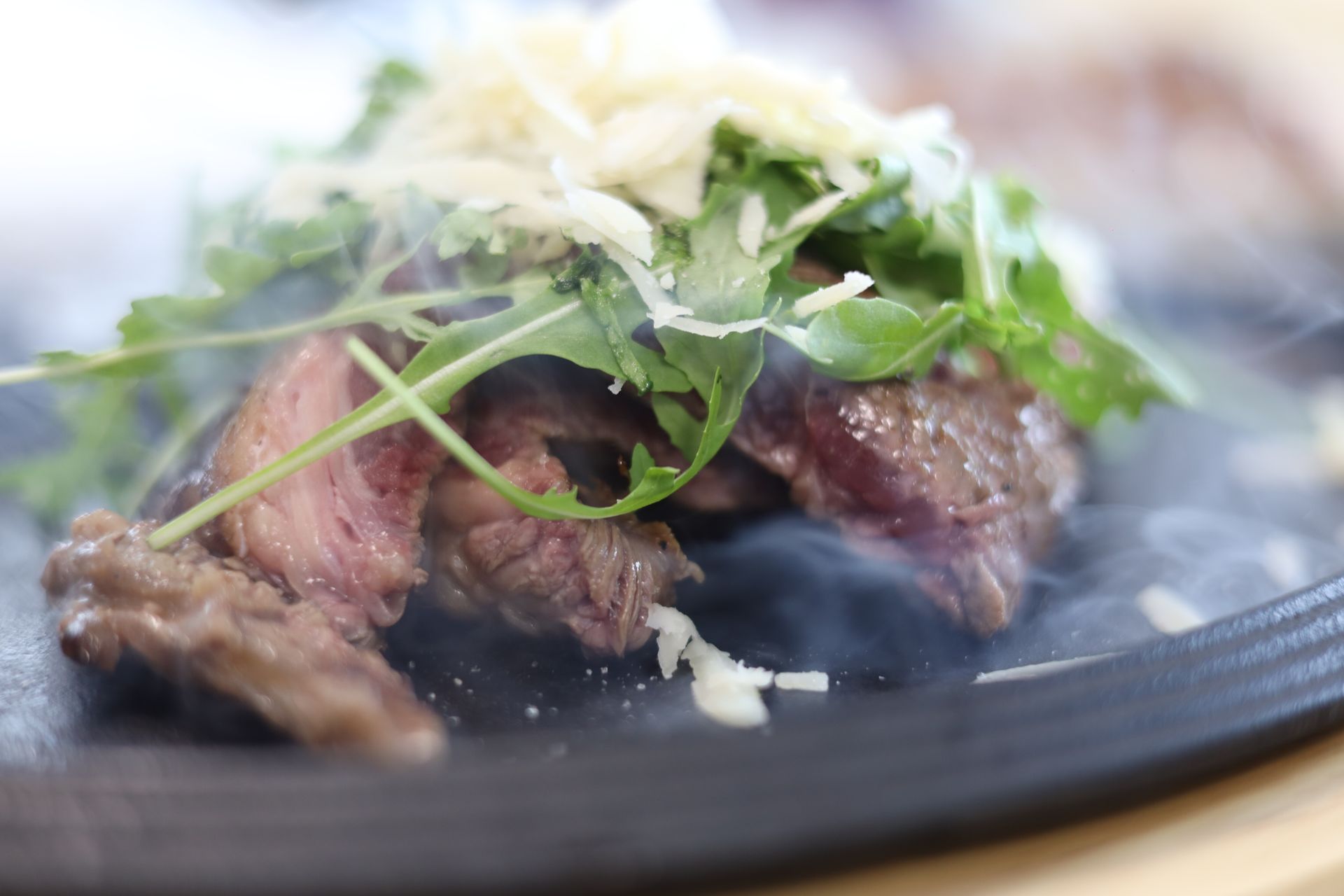 Bistecca tagliata a fette condita con rucola e formaggio grattugiato su un piatto scuro; si vede il fumo.
