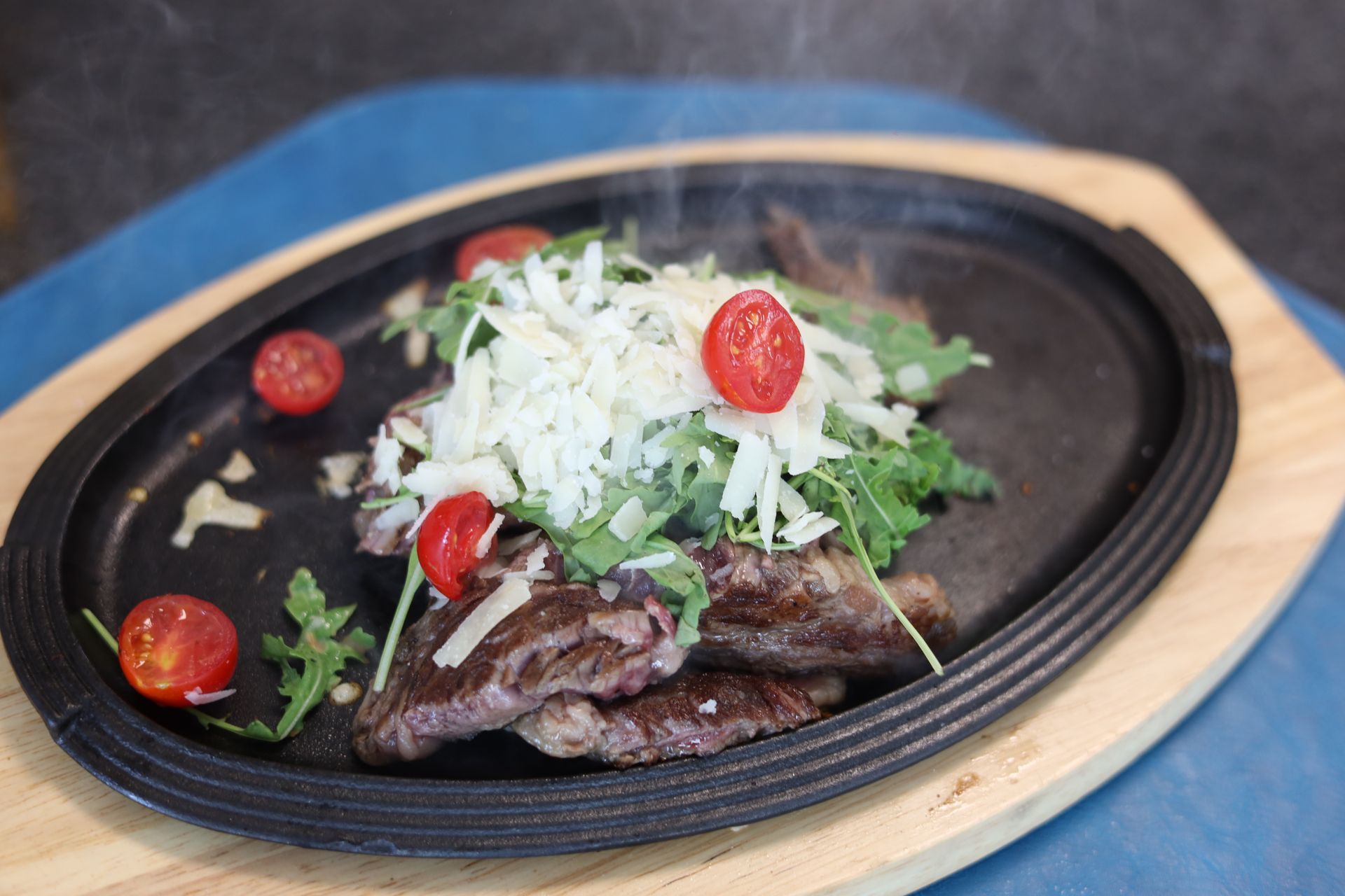 Bistecca sfrigolante con rucola, formaggio grattugiato e pomodorini su un piatto ovale nero.