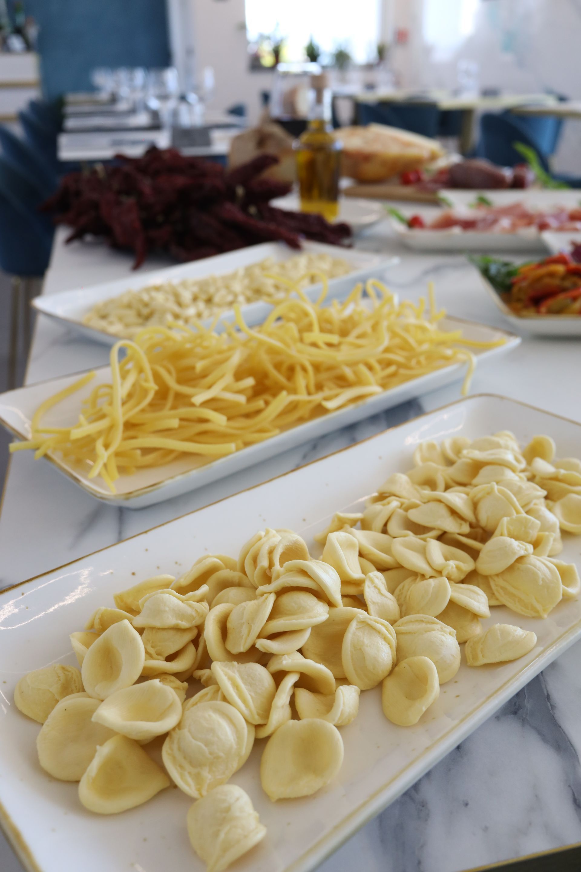 Forme di pasta esposte su piatti rettangolari bianchi. Altri ingredienti e l'ambiente della cucina sullo sfondo.