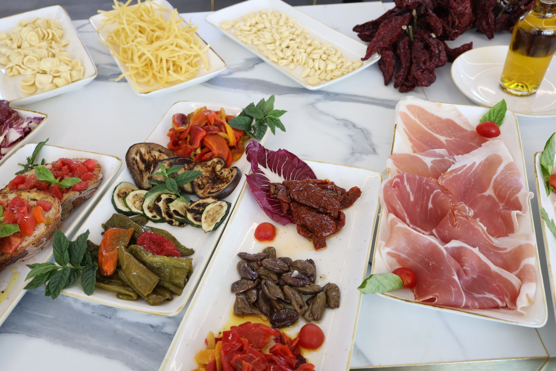 Un assortimento di antipasti italiani, tra cui pasta, salumi, verdure arrostite e pomodori secchi, sono esposti su una superficie di marmo bianco.