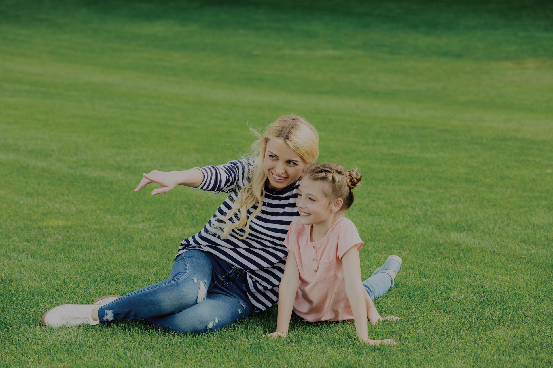 Woman and young girl sitting on green grass, smiling. The woman points toward the distance.