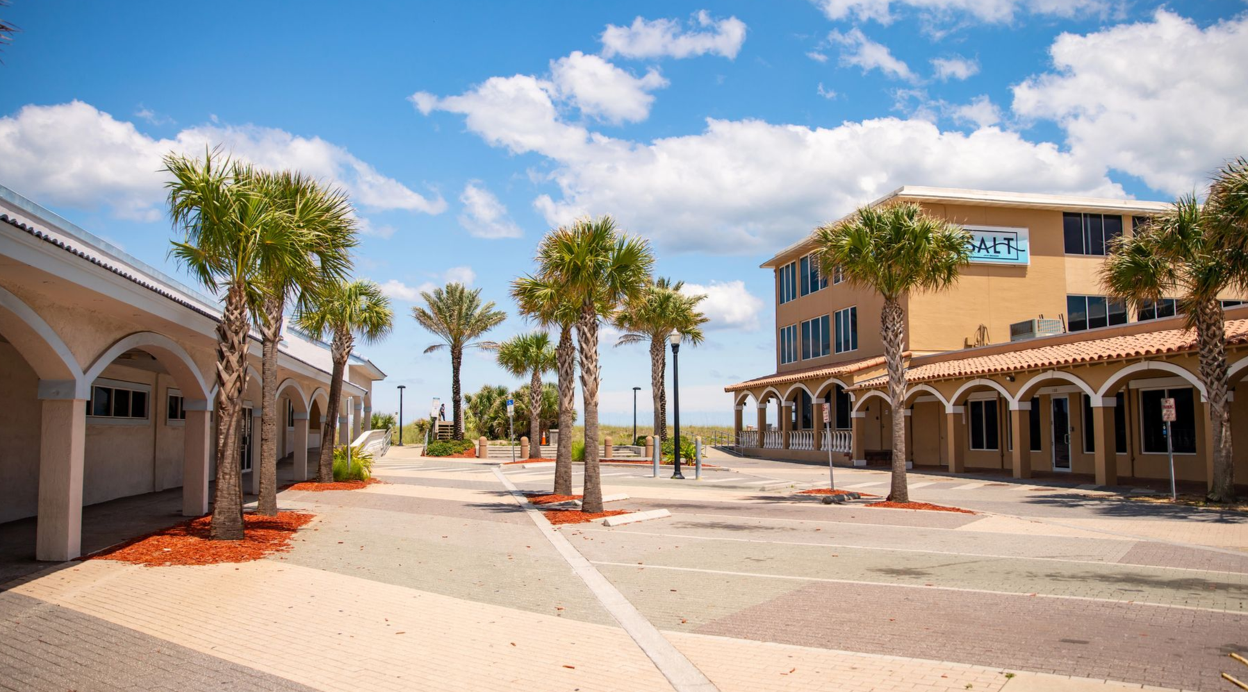 A pedestrian walkway at a beach, with palm trees, buildings, and a bright sky.