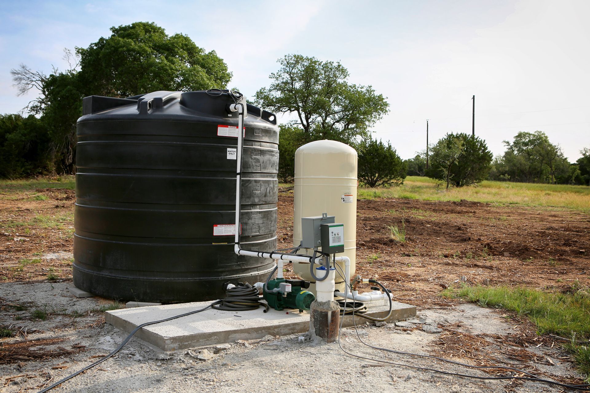 A black water storage tank and a beige pressure tank with associated equipment sit outdoors on a concrete pad in a field.