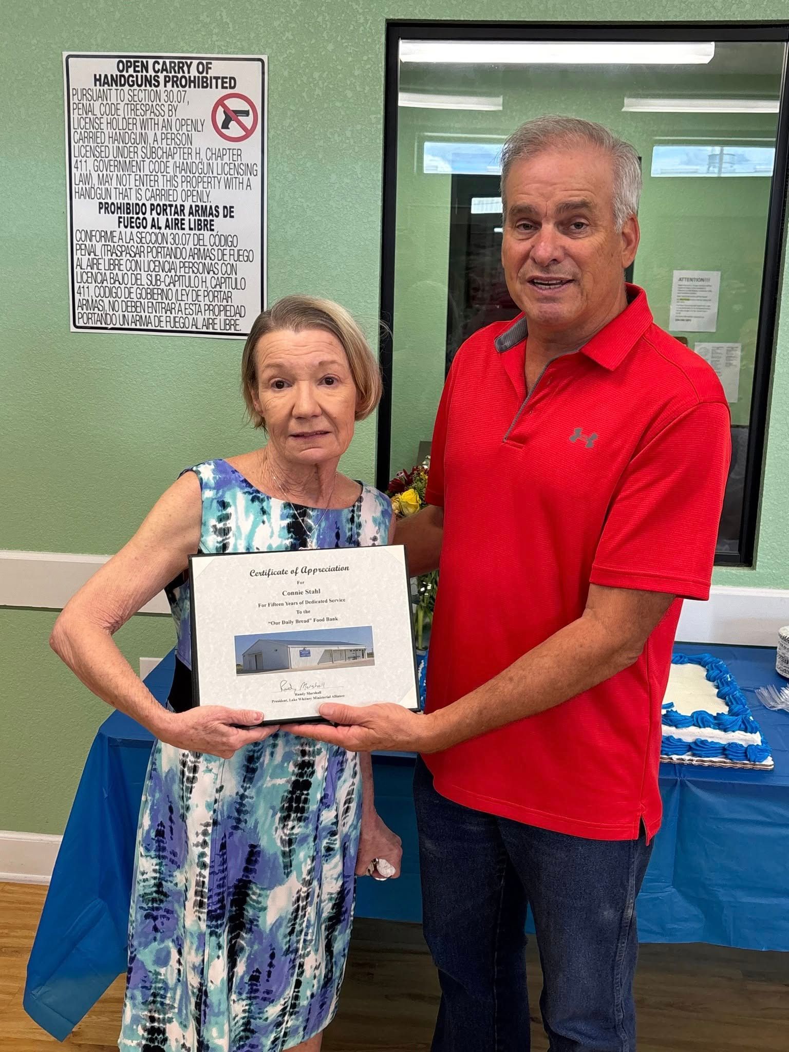 Two people holding a framed certificate; woman in blue dress, man in red shirt, inside setting.