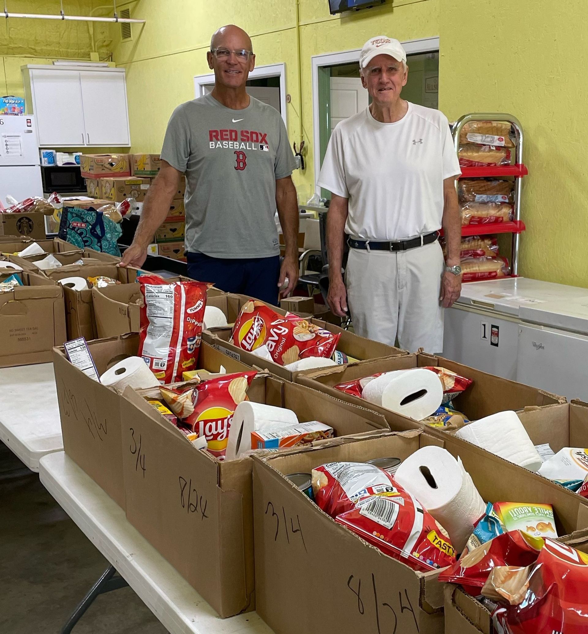 Two men are standing in front of a table filled with boxes of food.