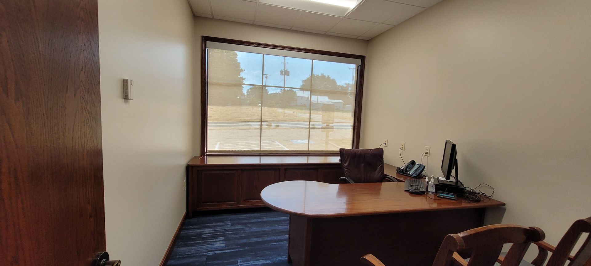 Office with a desk, window, chair, and dark-stained door. The walls are beige, the floor is dark blue.