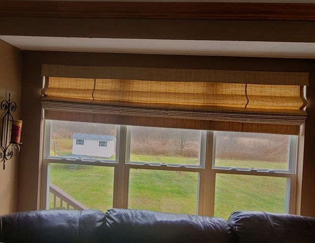 Window with light brown woven shade, showing a grassy yard and small shed. Brown trim and dark couch visible.