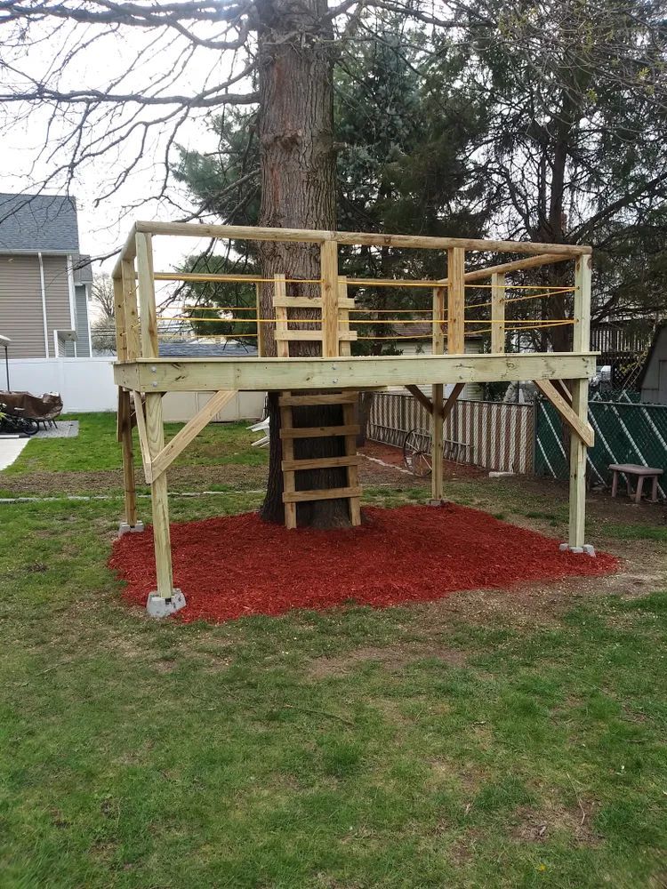 A tree house with a deck built around a tree in a backyard